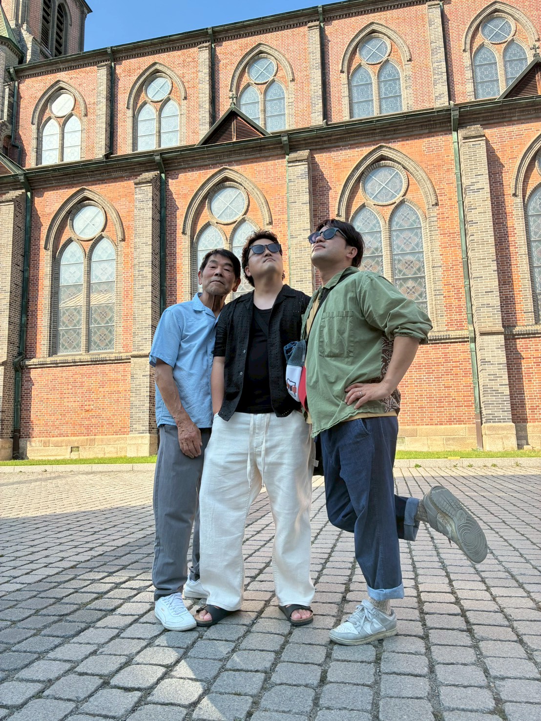 LINE_ALBUM_20260610_260622_10 - 滋賀で自然素材・木の家の注文住宅を建てる工務店|株式会社スムース Three men stand on a cobblestone plaza in front of a red brick church with tall arched windows, looking up at the sky.
