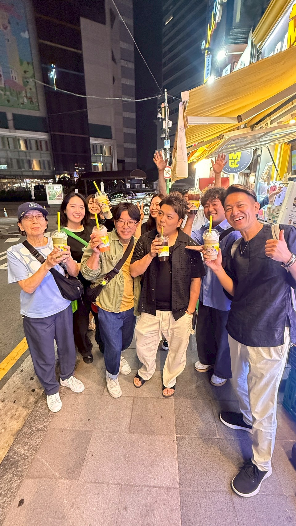 LINE_ALBUM_20260610_260622_5 - 滋賀で自然素材・木の家の注文住宅を建てる工務店|株式会社スムース Group of eight friends posing on a city sidewalk at night, holding iced drinks with yellow straws and smiling at the camera in front of a yellow storefront awning.