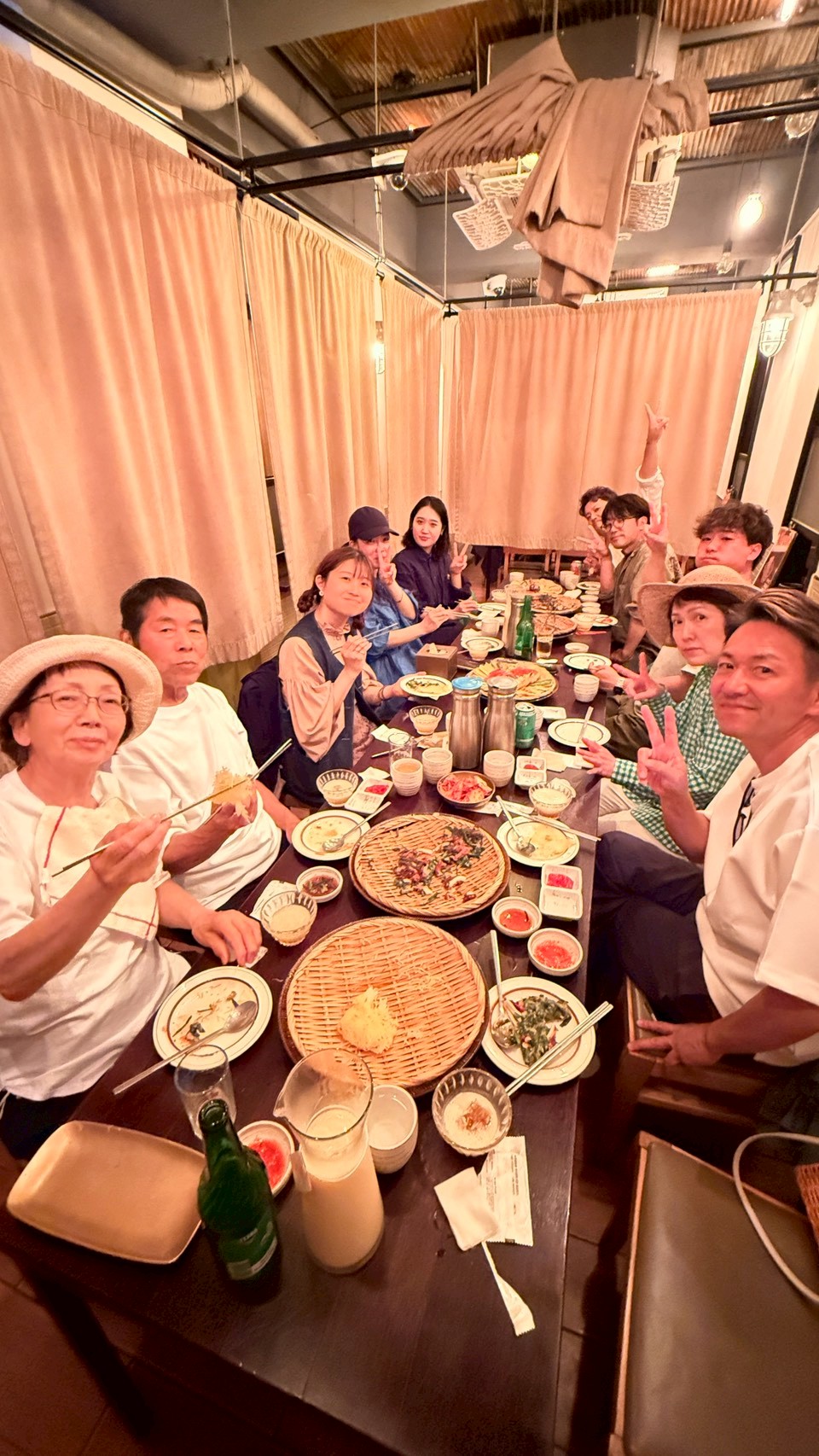 LINE_ALBUM_2026611社員旅行2日目_260622_10 - 滋賀で自然素材・木の家の注文住宅を建てる工務店|株式会社スムース A group of friends around a long table in a cozy restaurant, smiling and flashing peace signs as they share plates and bowls of food.