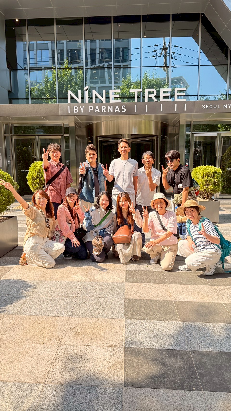 LINE_ALBUM_DAY3_260622_3 - 滋賀で自然素材・木の家の注文住宅を建てる工務店|株式会社スムース Group of about a dozen people posing for a photo in front of a modern glass hotel entrance (Nine Tree by Parnas, Seoul), many showing peace signs on a sunny day.