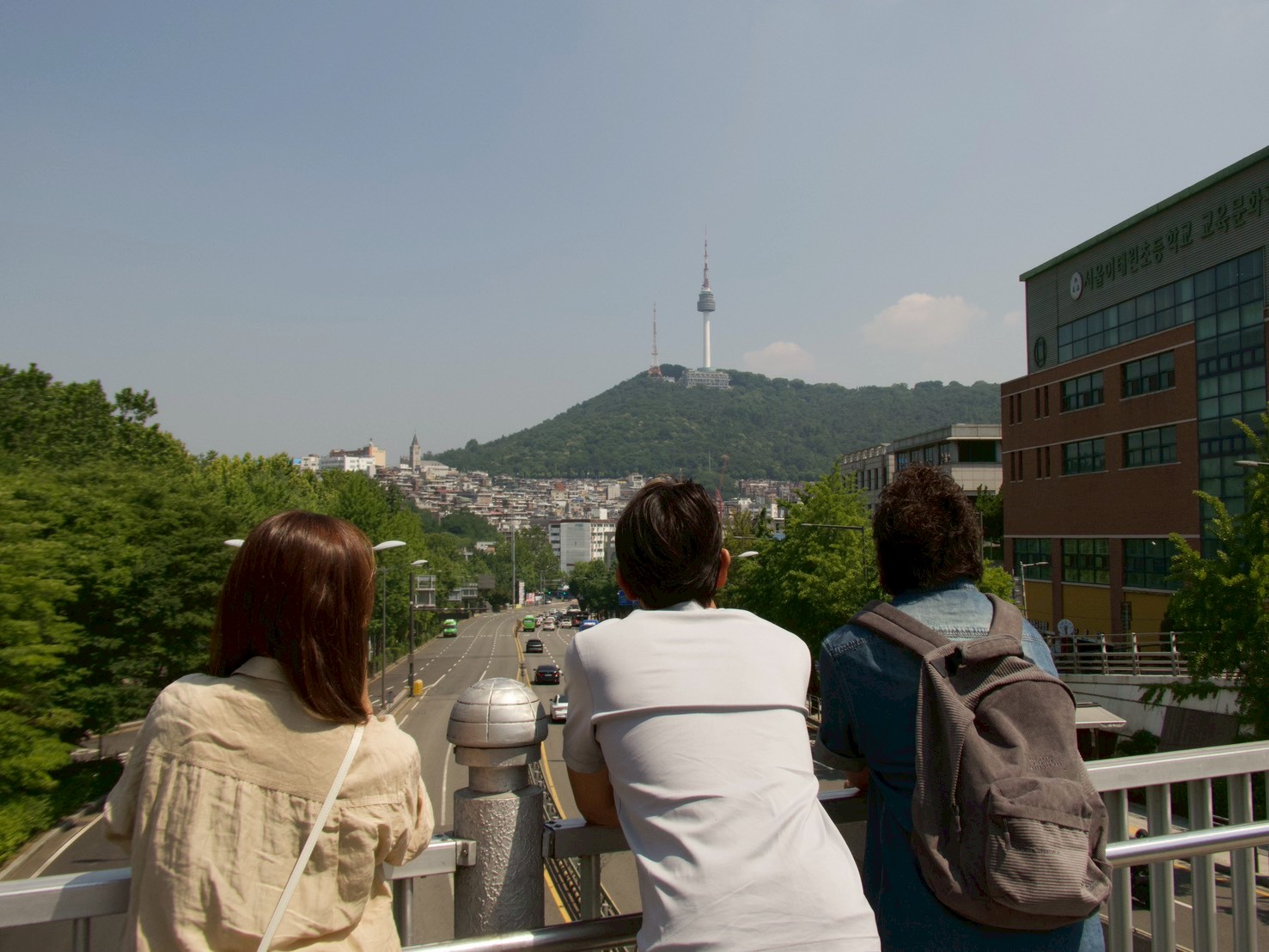 LINE_ALBUM_DAY3_260622_8 - 滋賀で自然素材・木の家の注文住宅を建てる工務店|株式会社スムース Three people stand at a railing on a bridge, facing a cityscape with a green hill and a tall tower in the distance.