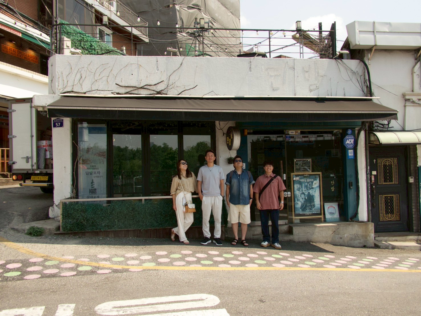 LINE_ALBUM_DAY3_260622_9 - 滋賀で自然素材・木の家の注文住宅を建てる工務店|株式会社スムース Four casually dressed people pose in front of a small storefront with a black awning on a sunny street outside a shop door and windows.