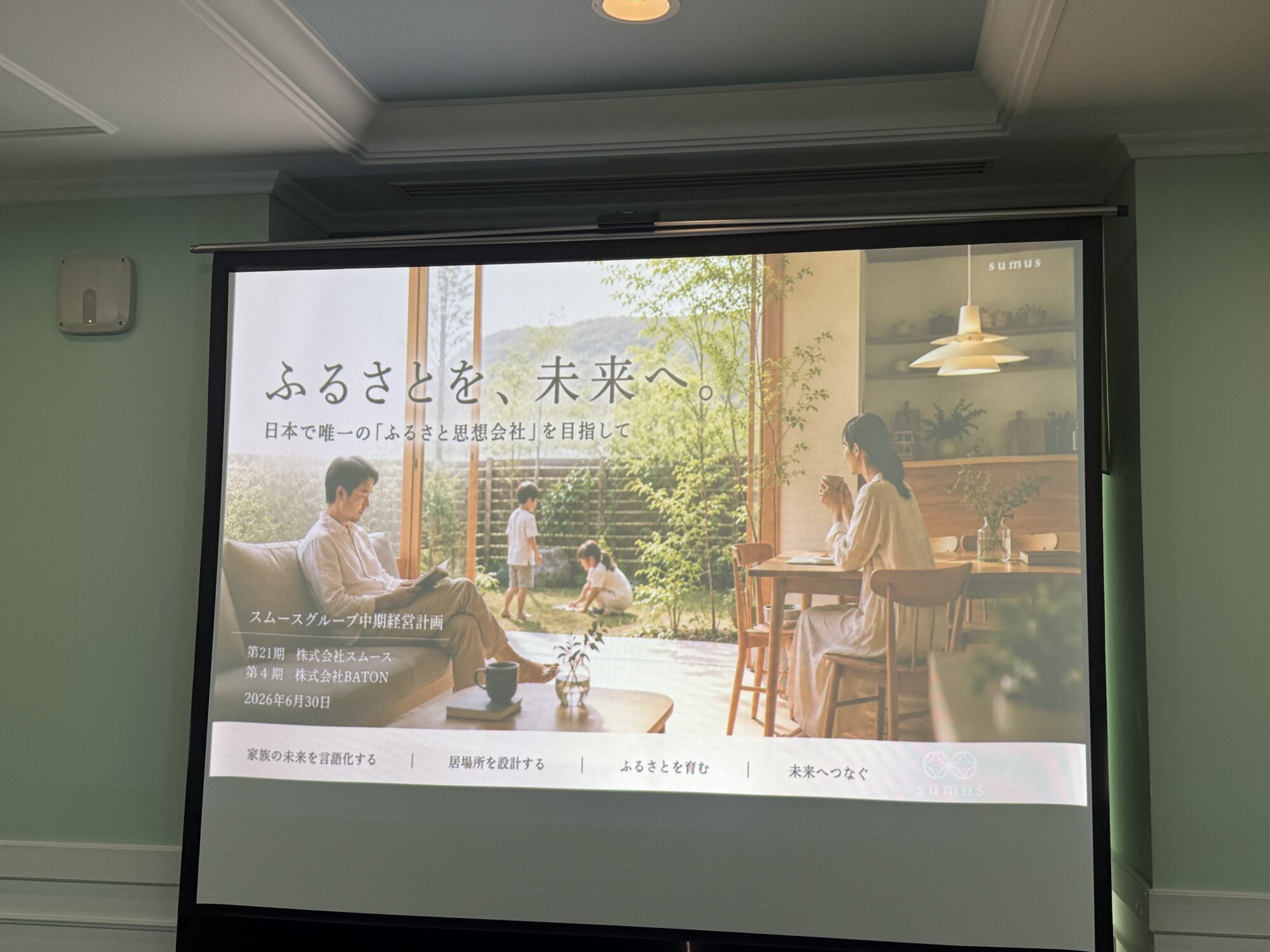 Large conference room screen showing a Japanese ad with people at a cafe garden scene; greenery and text visible on screen edge.