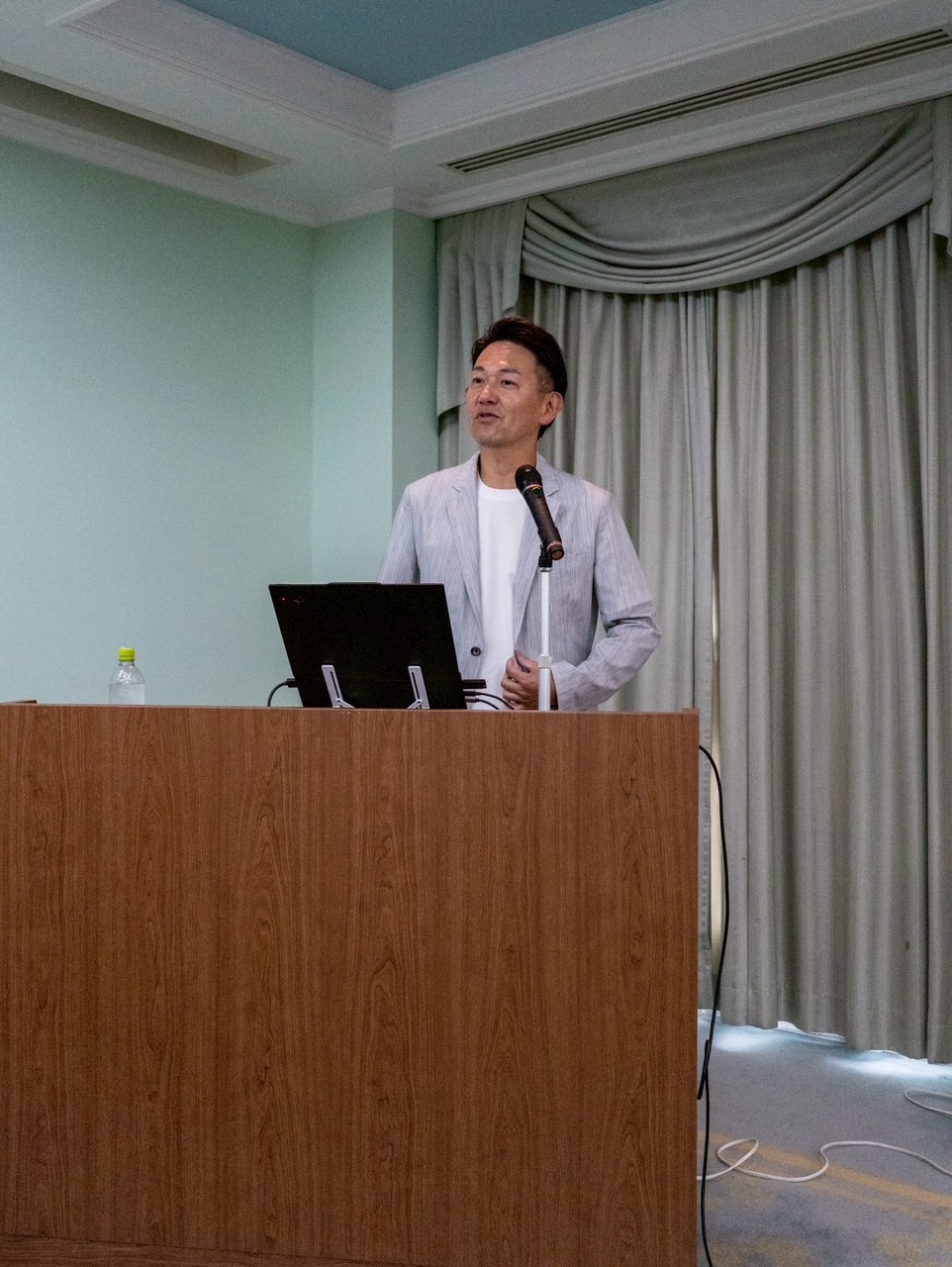Man in a light blazer speaks at a wooden lectern with a laptop and microphone in a conference room.