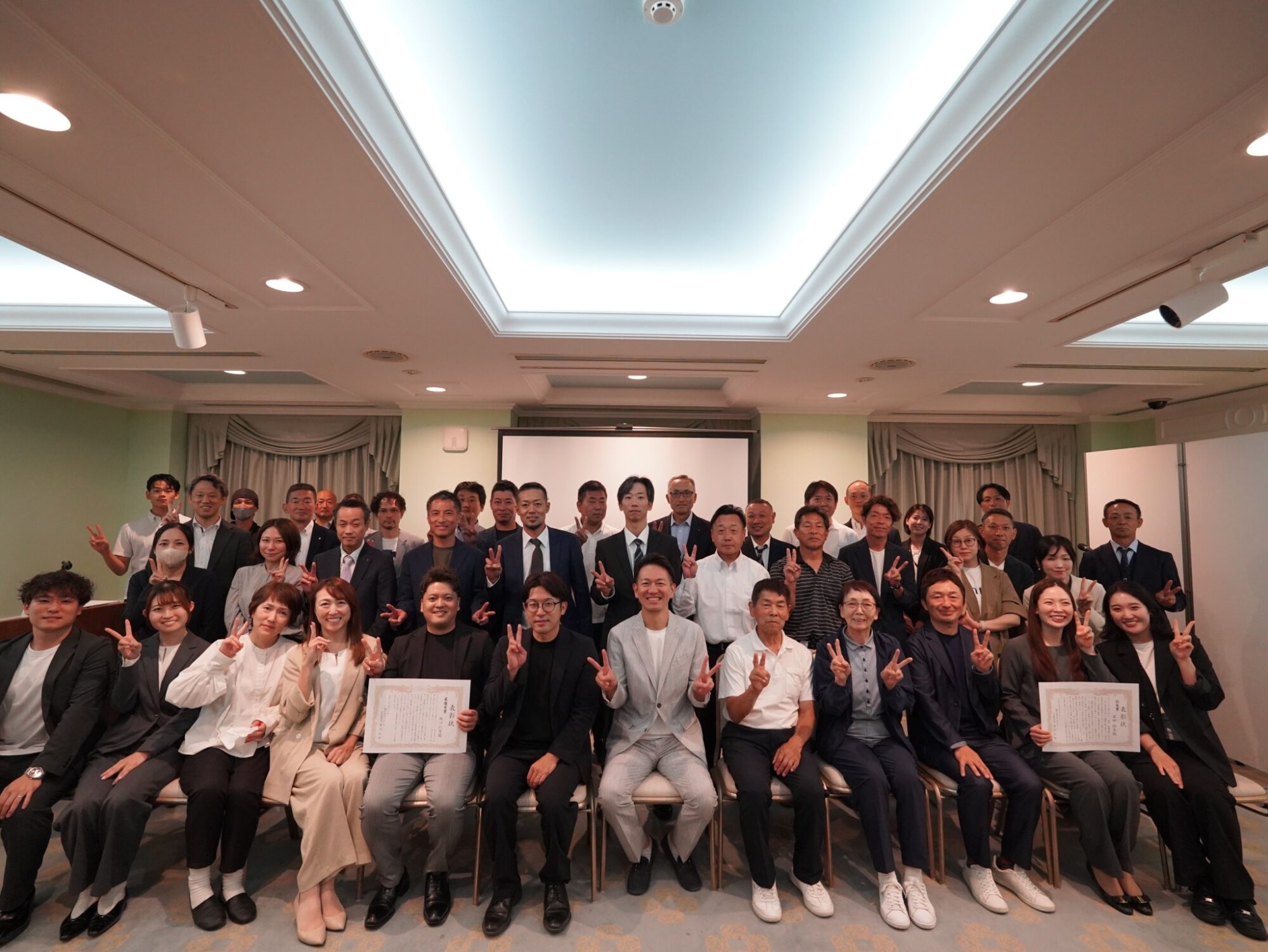 Large group of people in business attire posing for a group photo indoors, many making peace signs, some holding certificates.