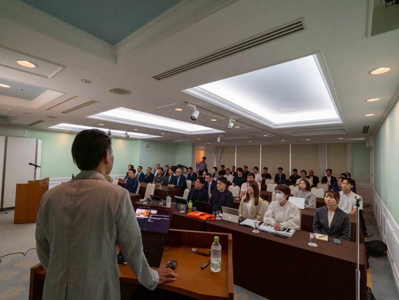 Speaker at a wooden podium presenting to a large group in a conference room.