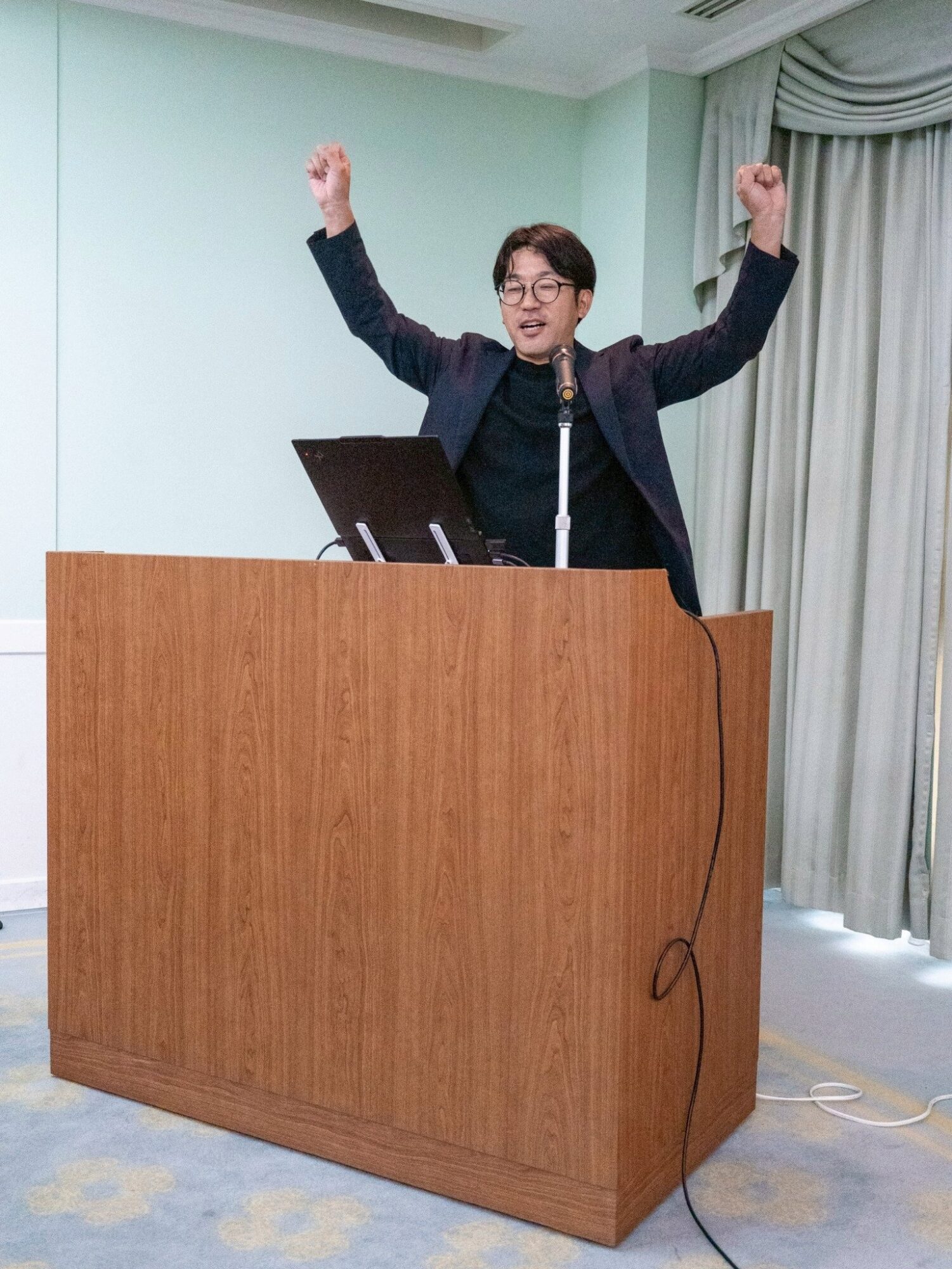 Man at a wooden podium raises both arms in victory, speaking into a microphone in a conference room