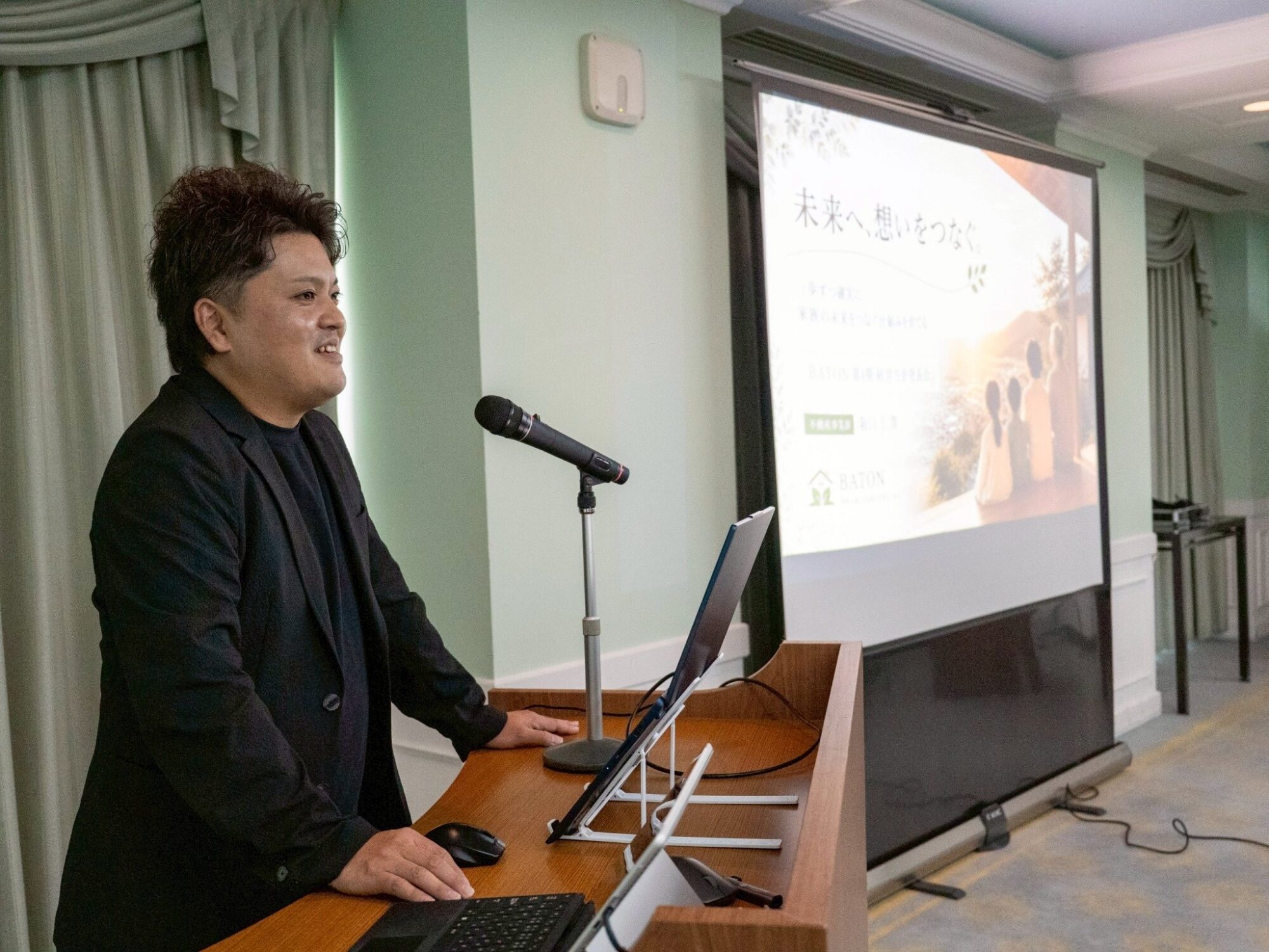 Man in a black blazer giving a presentation at a lectern with a microphone, projection screen showing slides behind him.