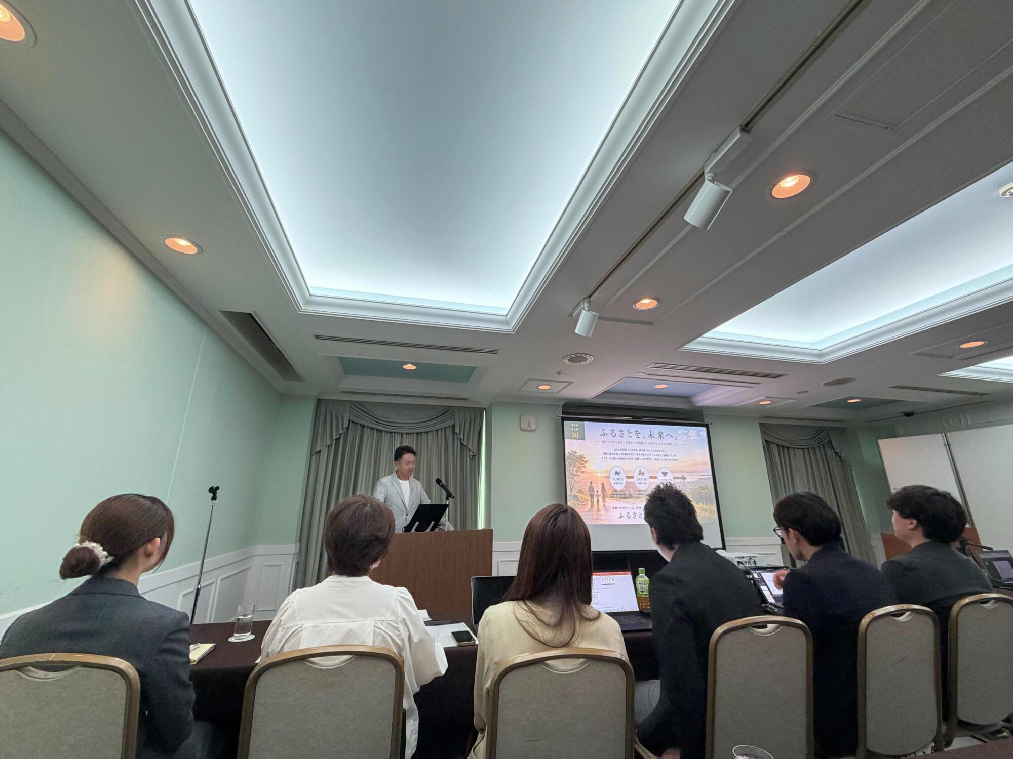 Man at a podium giving a presentation in a conference room as the audience watches; a slide with Japanese text is displayed on a screen behind him.