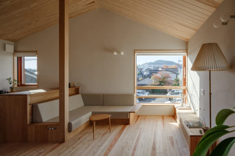 Bright attic living room with wood-paneled walls, beige L-shaped sofa, round wooden coffee table, and a large window overlooking a town.