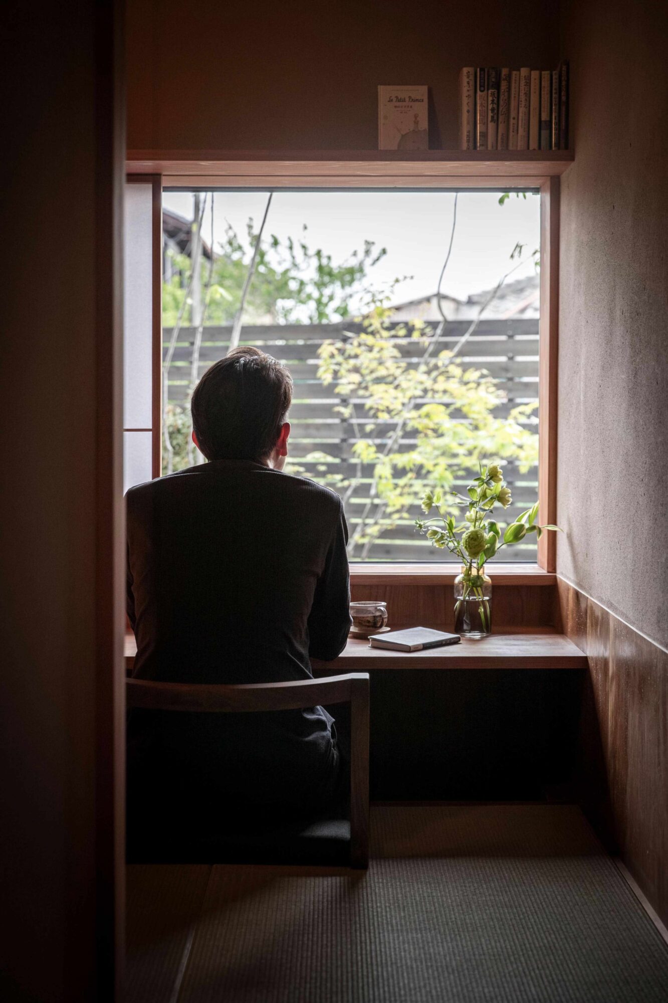 _X1I0727-br 1 - 滋賀で自然素材・木の家の注文住宅を建てる工務店|株式会社スムース Person sits with back to the camera at a wooden desk by a large window, gazing outward at greenery; notebook and glass on the desk.