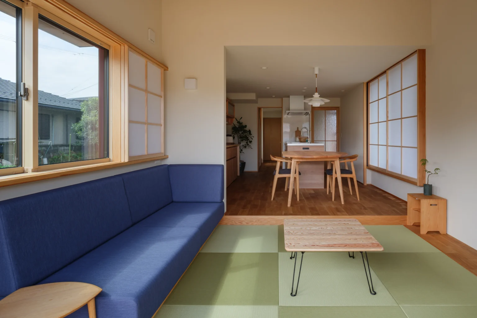 251014_049-1536x1024 - 滋賀で自然素材・木の家の注文住宅を建てる工務店|株式会社スムース Open-plan living room with a blue sofa on tatami mats, a wooden coffee table, and sliding shoji screens; dining area and kitchen in the back.