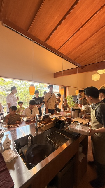 Open kitchen and dining area with a wooden ceiling; people gather around a stainless counter, some eating, a server attends to guests.