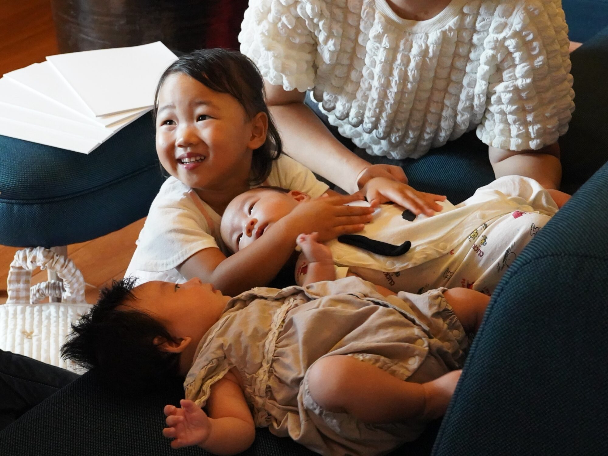 Smiling toddler cuddling a baby on a dark blue sofa, with an adult in a white textured sweater nearby and white papers on the armrest