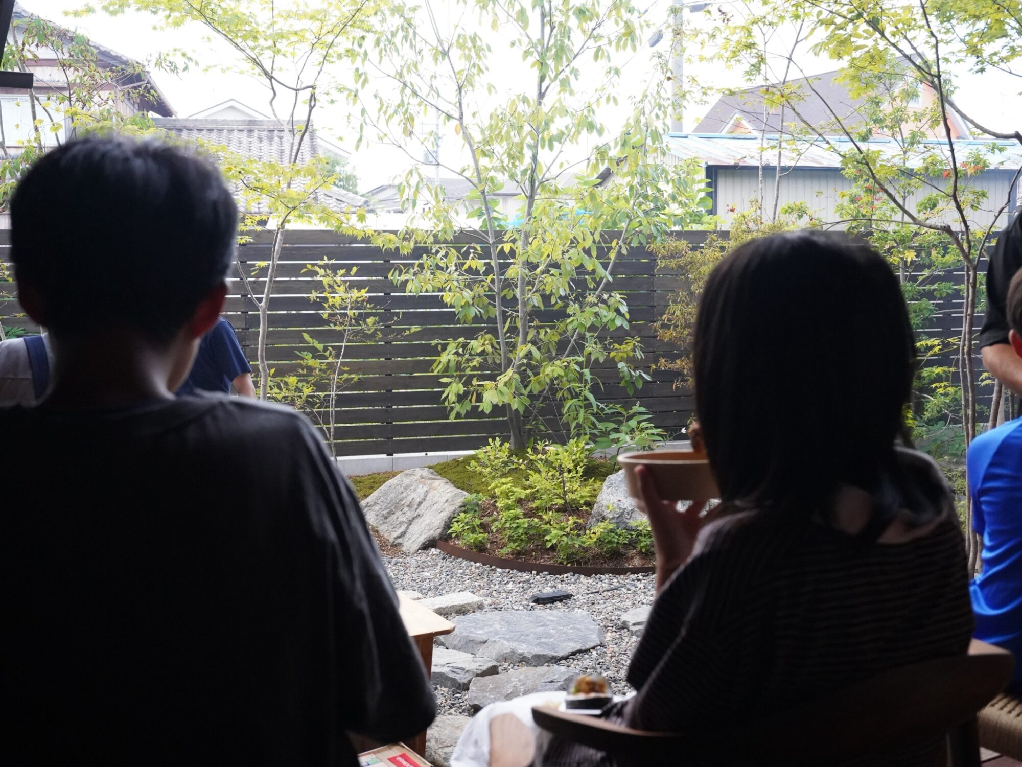 People seated with their backs to the camera in a backyard garden, trees above and a dark fence in the background, with small plates of food on a table nearby