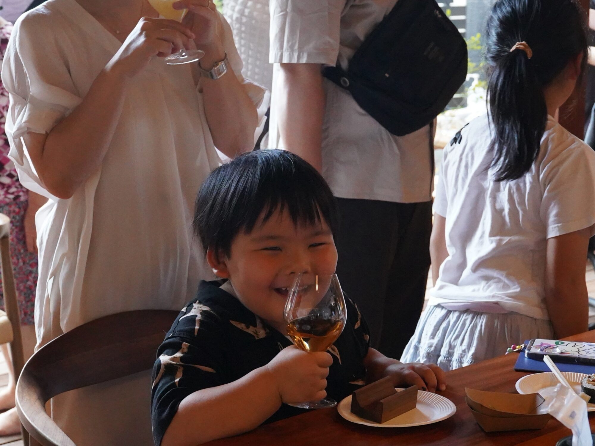 Smiling young boy at a table holding a wine glass with desserts nearby, surrounded by adults in white shirts.