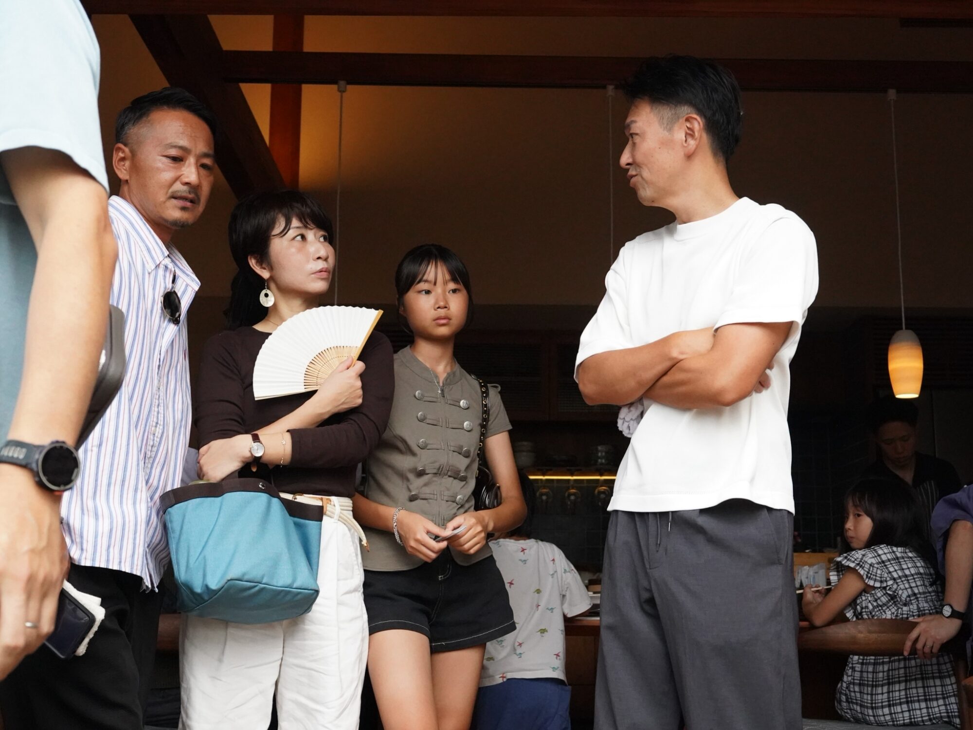Group of people chatting in a dim cafe; a woman in black holds a white folding fan while others listen.