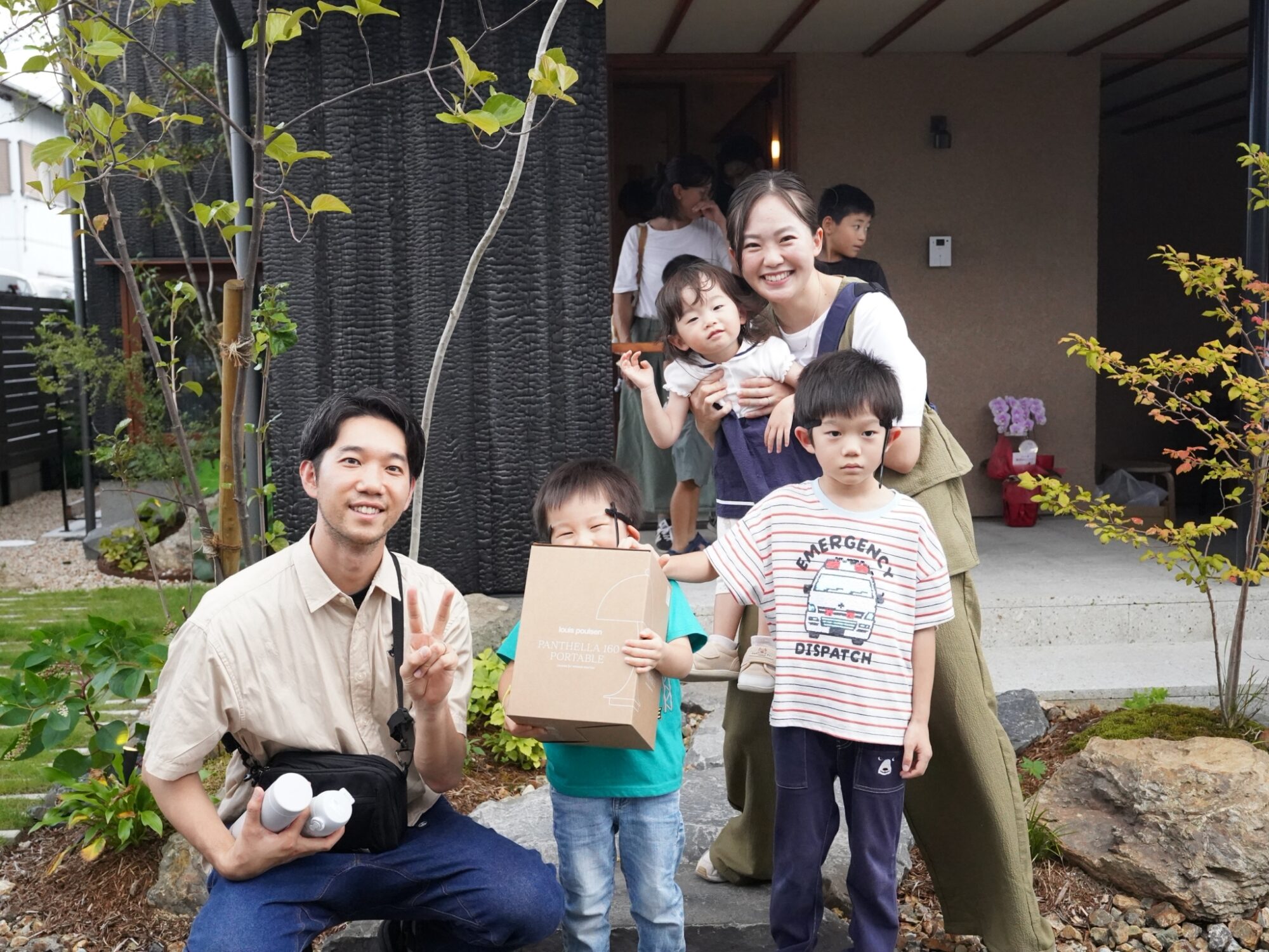 Smiling family and friends pose for a group photo outdoors on a garden path, with two children in front holding a gift box or bag.