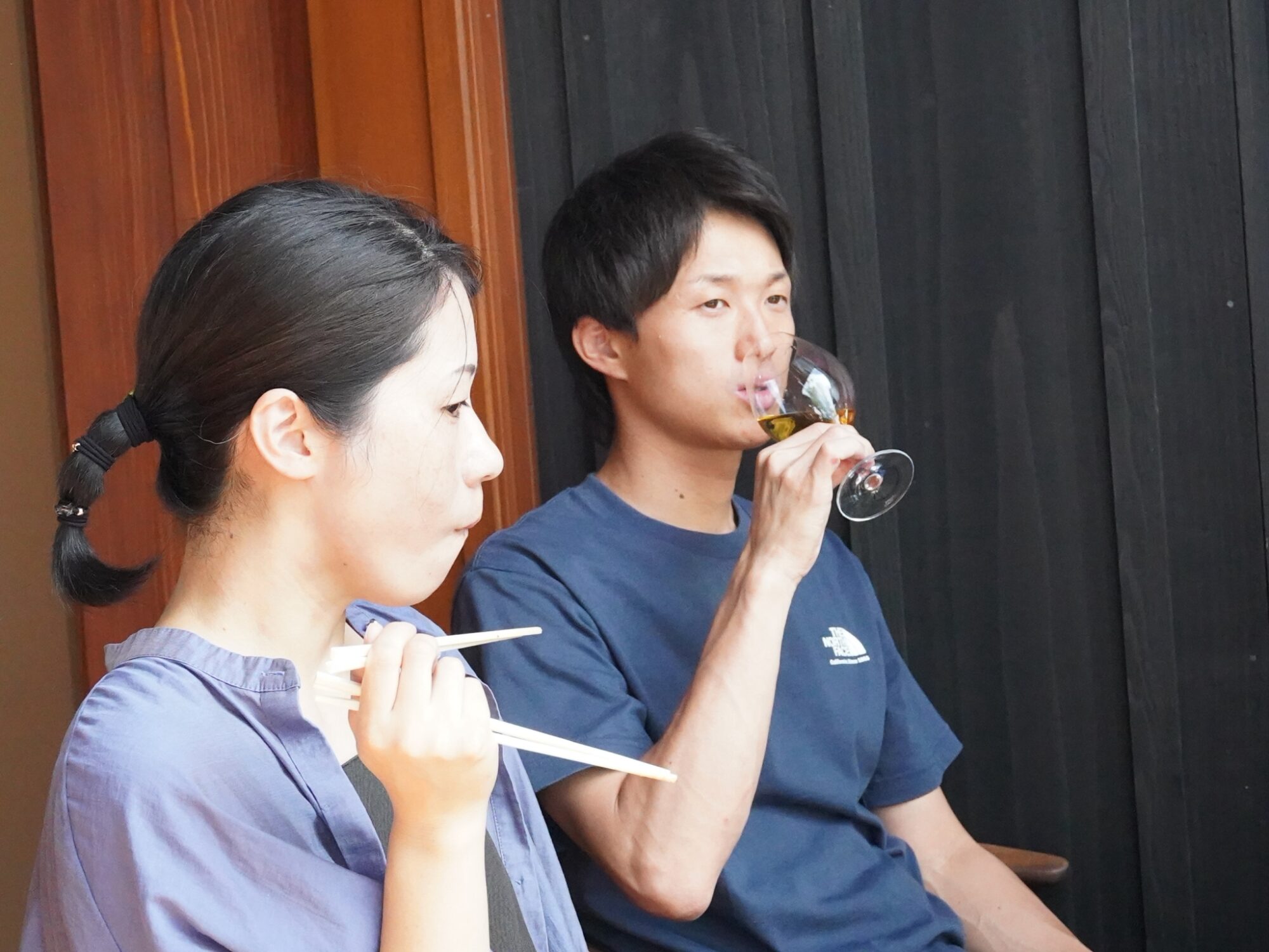 Two people seated indoors/outdoors near wooden panels: a woman in a purple top holds chopsticks, a man in a blue shirt sips white wine from a glass.