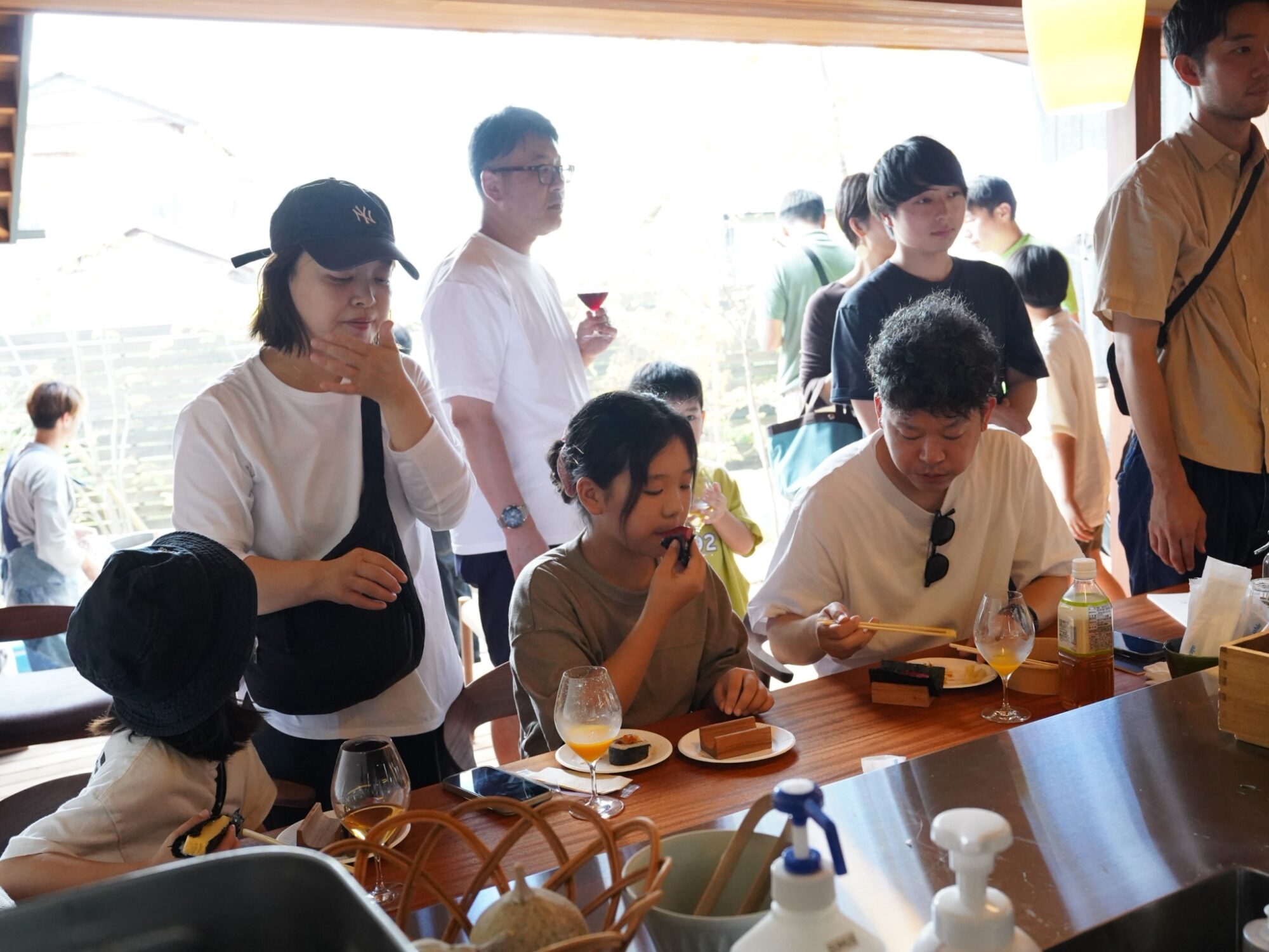 Group of people around a wooden table with plates and drinks in a bright dining room, some standing and others seated, social gathering.