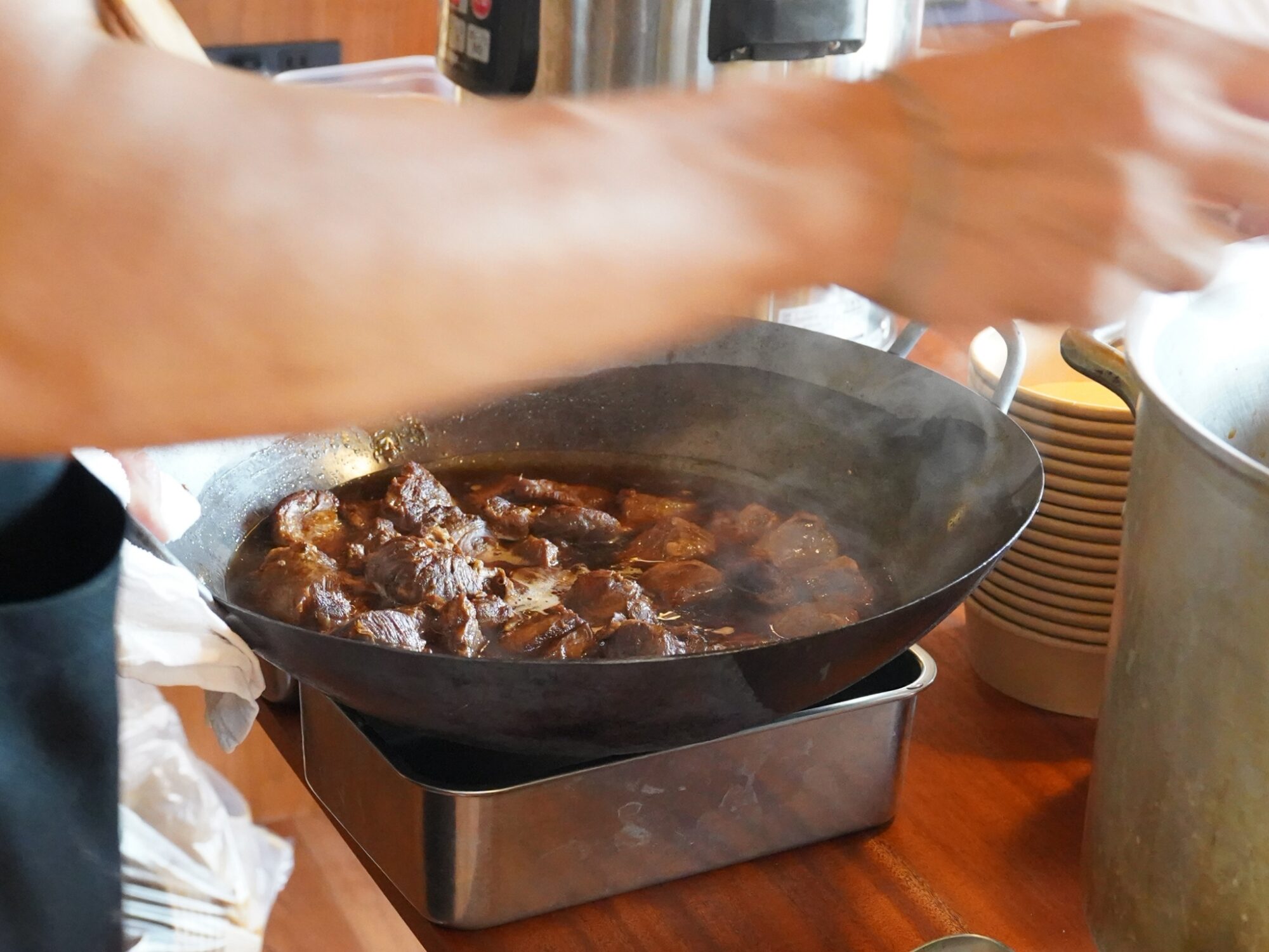 Chunks of browned meat simmering in a large skillet with steam rising from the sauce near a kitchen stove.