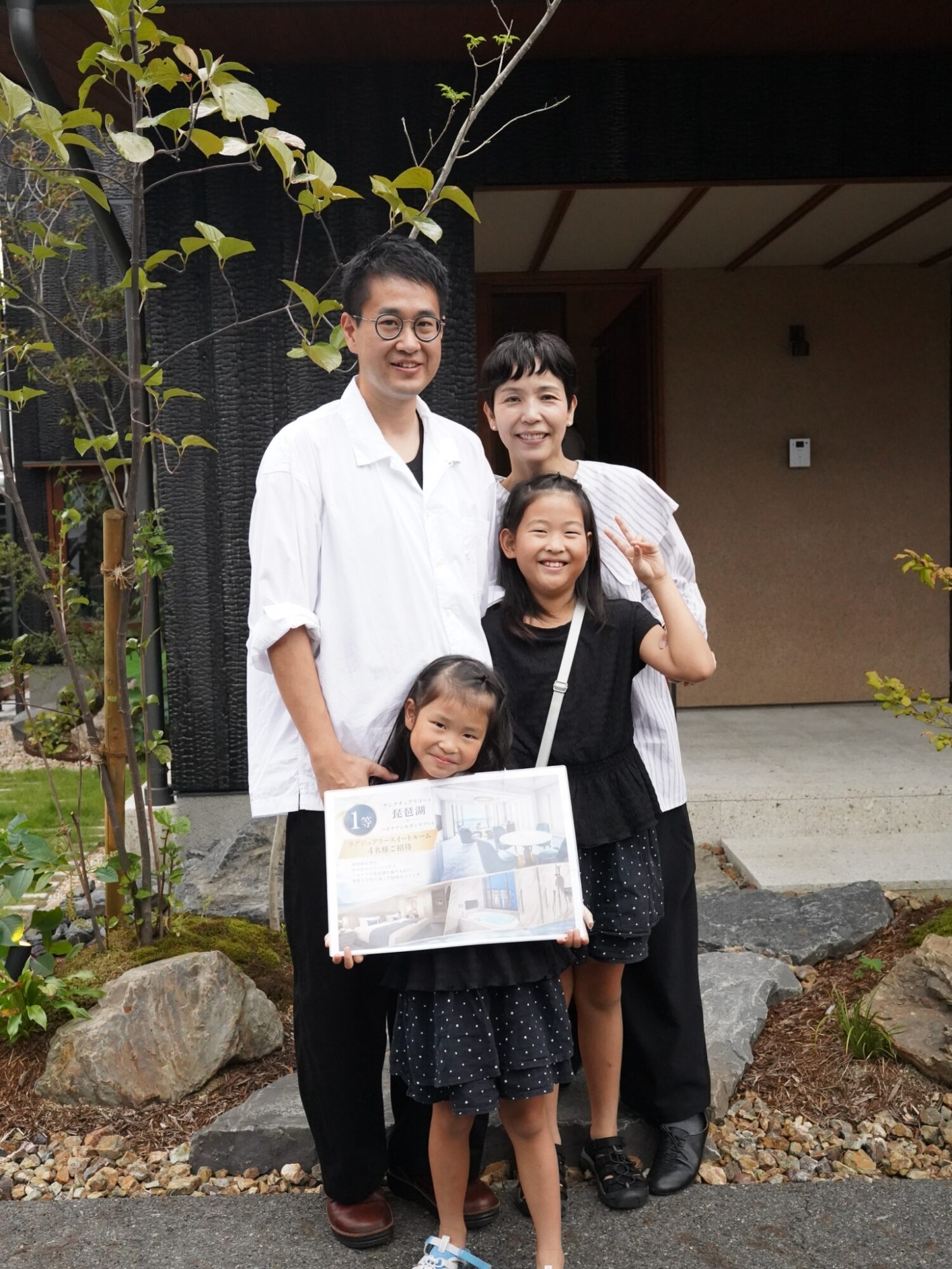 Smiling family of four posing outside a house, with a young girl in front holding a large poster posterboard.