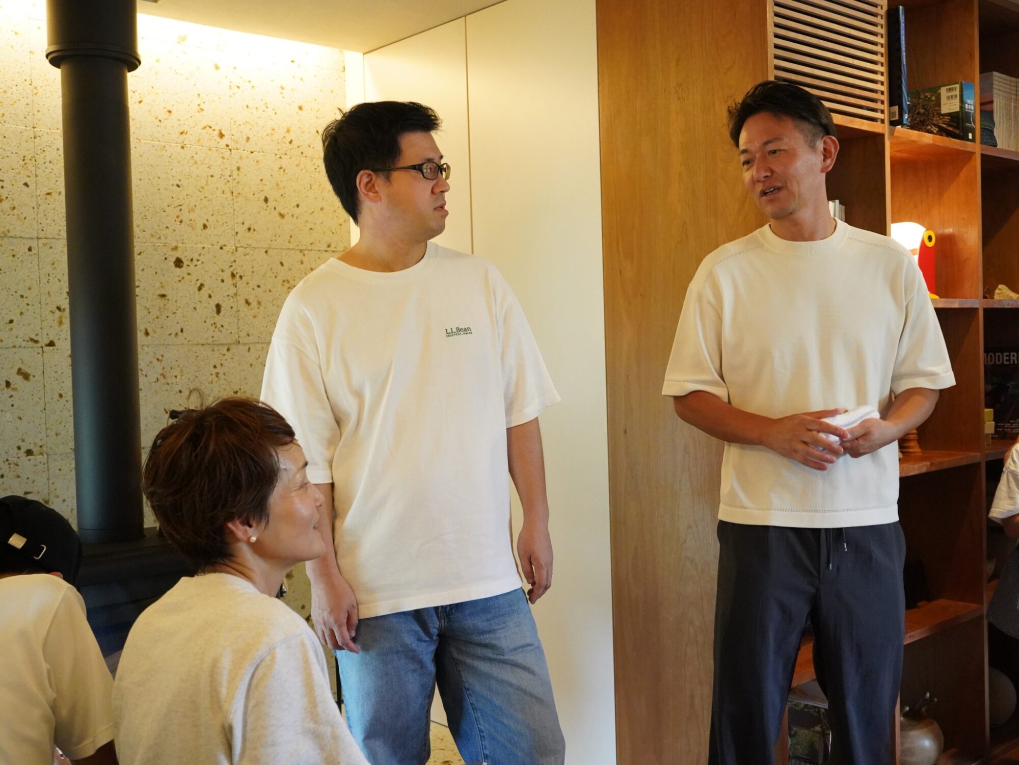 Two men in white T-shirts converse in a warm wooden-room setting while others sit nearby.