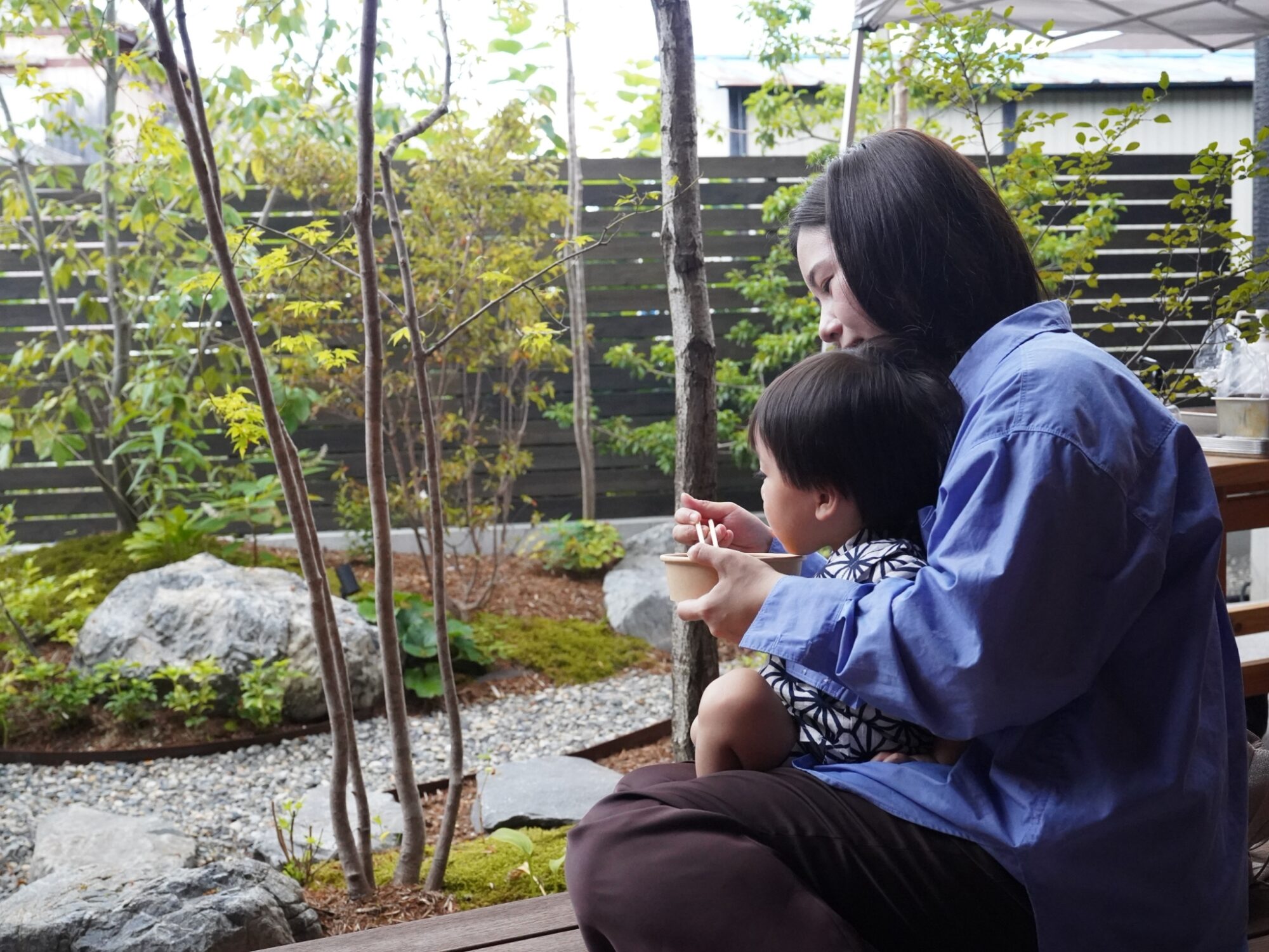Woman in a blue shirt sits on a bench with a young child, sharing food in a serene garden with rocks and green plants.