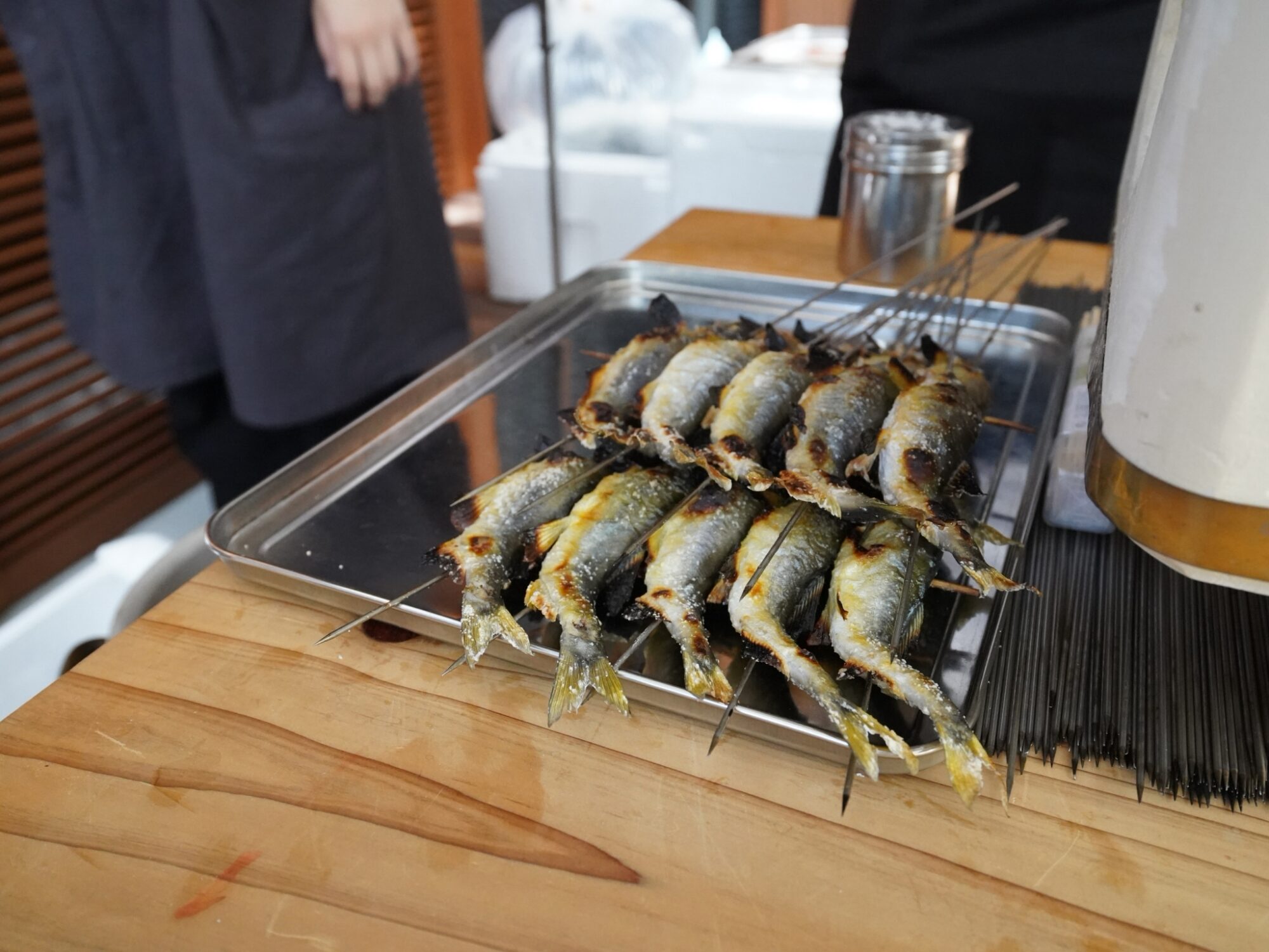 Skewered grilled small fish arranged on a metal tray over a wooden table in a kitchen setting.