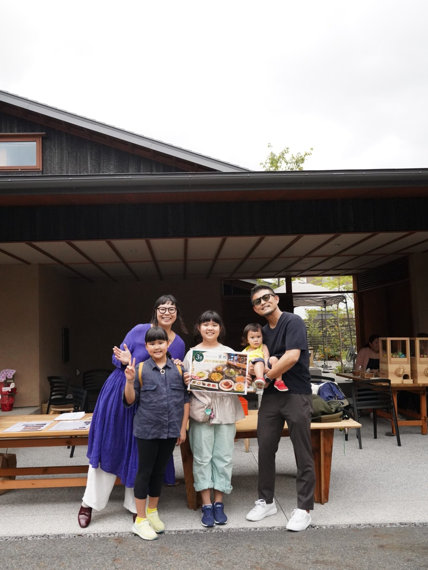 Group of family members outdoors, smiling, holding a large menu-board in front of a house with wooden furniture.