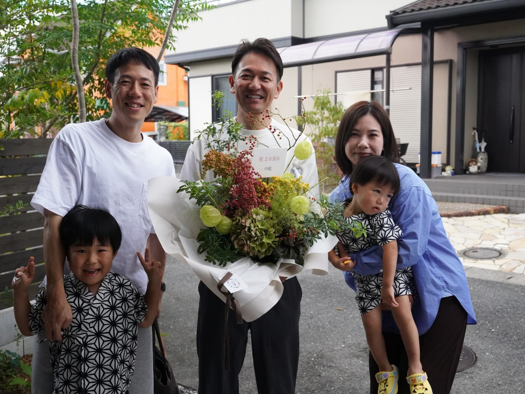 LINE_ALBUM_20周年_260926_6 - 滋賀で自然素材・木の家の注文住宅を建てる工務店|株式会社スムース Three adults and two children pose outdoors with a large flower bouquet, celebrating a 20th anniversary.
