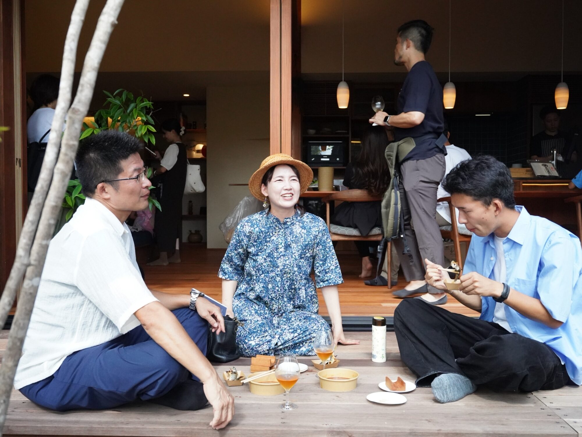 Group of people enjoying drinks and snacks on a wooden patio; woman in a straw hat smiles at center.